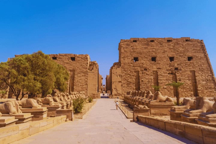 Pathway lined with sphinx statues and ancient ruins.